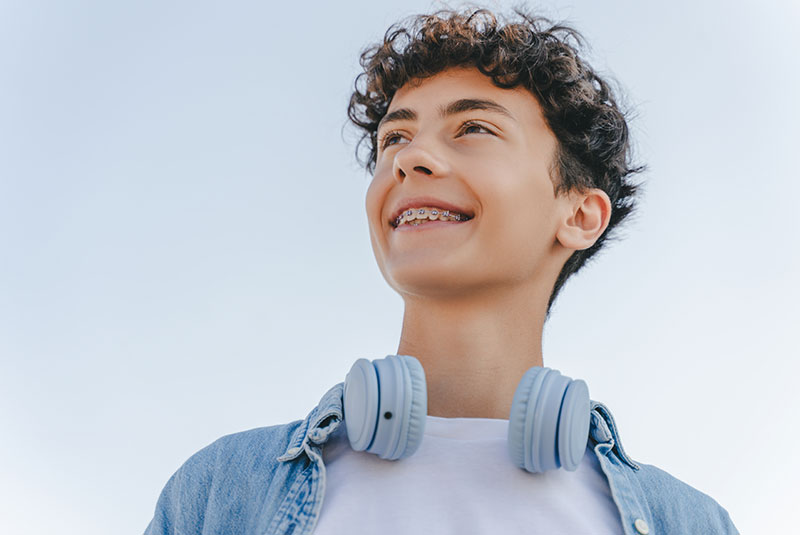 smiling curly haired teenager with braces wearing headphones looking away standing on the street