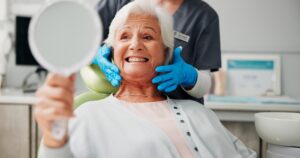 An older woman with white hair grins while holding a hand mirror in a dental chair. A dental professional wearing blue gloves and a gray uniform stands behind her, holding her chin to examine her teeth.