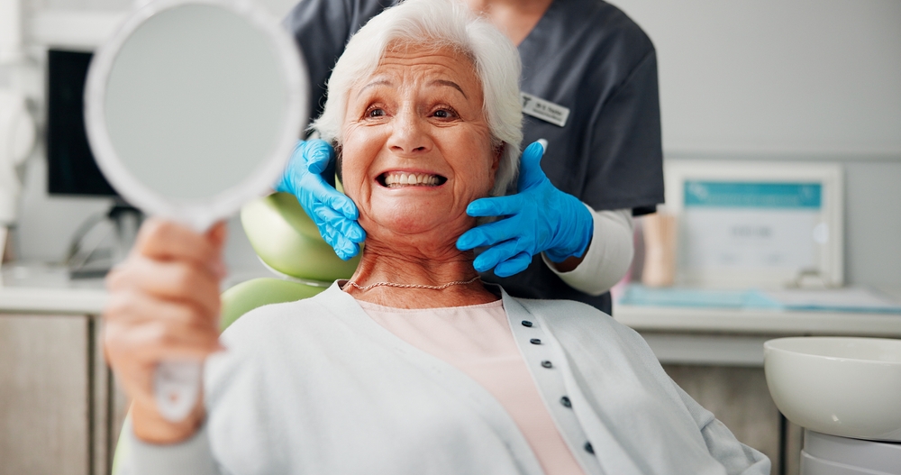An older woman with white hair grins while holding a hand mirror in a dental chair. A dental professional wearing blue gloves and a gray uniform stands behind her, holding her chin to examine her teeth.