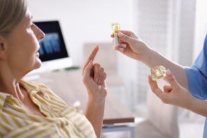 A dentist holds a model of a dental implant and crown for a patient to inspect. The patient, wearing a yellow striped shirt, listens attentively in a bright, modern office with a blurred dental X-ray visible on a monitor in the background.